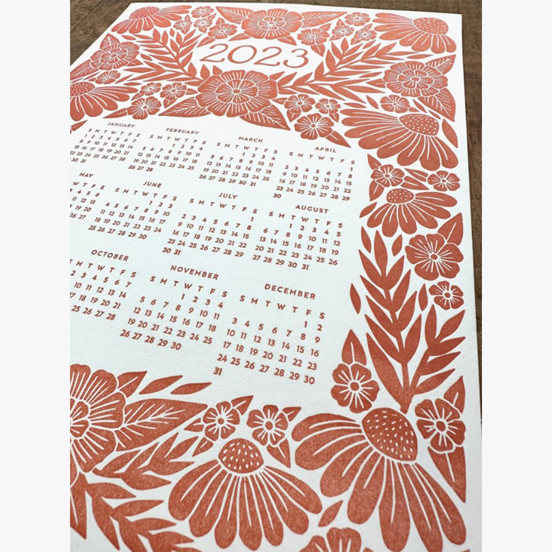 Our Red Floral Letterpress 2023 Calendar showcases a detailed floral graphic design that's letterpress printed with red ink on a thick museum, off-white board. The number 2023 is the headline on the upper third of the page, and the annual calendar (January through December,) is featured on the mid and lower third of the page. The 2023 calendar and 2023 headline are both surrounded by the floral graphic design. This photo shows a close-up of most of the one-page calendar.